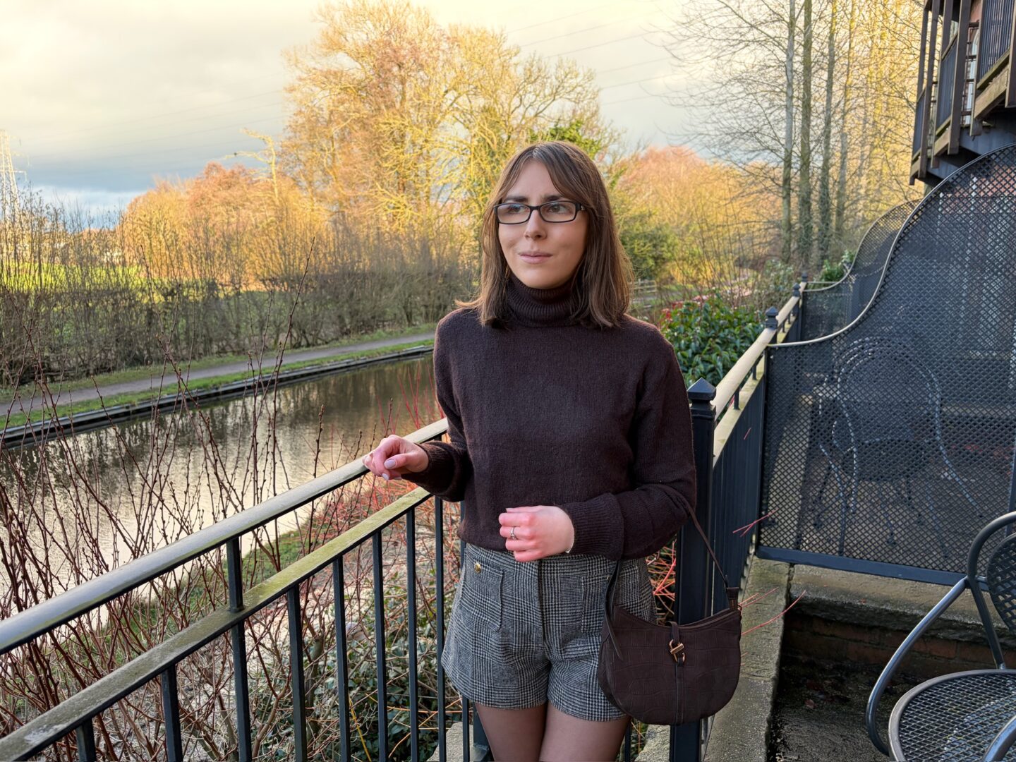 Elin standing with her right hand resting on a black railing that lines an outdoor terrace beside a canal. She’s looking away from the camera to her right with a neutral expression on her face. The water reflects the warm light and the trees and shrubs that border one side.