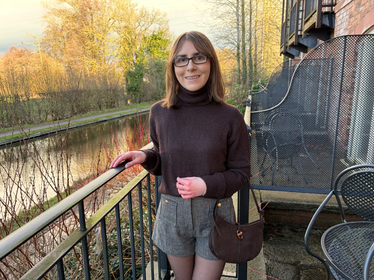 Elin standing on an outdoor terrace beside a canal. Her right hand rests on a black railing and she’s smiling broadly at the camera. She wears a brown high neck jumper with grey and brown check tailored shorts and brown tights. A brown shoulder bag with gold hardware and a croc style design hangs on the left arm across her hip. Her shoulder length brown hair is worn straight.