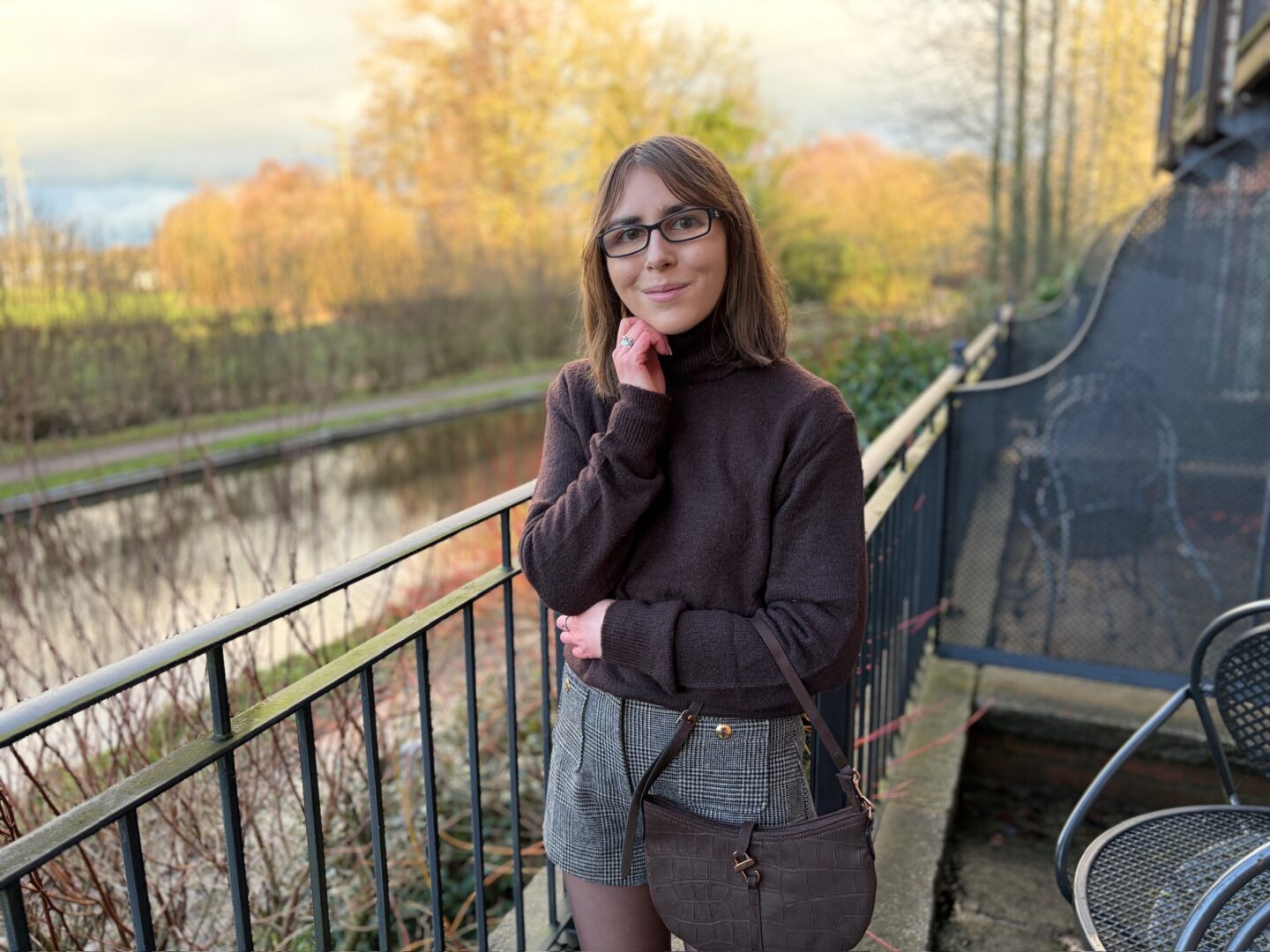 Elin standing on an outdoor terrace beside a canal. She wears a brown high neck jumper with grey and brown check shorts and light brown tights. She’s smiling a no teeth smile at the camera with her right hand resting under her chin and her left arm folded across her body. A brown bag hangs in the crook of her left elbow. The water beside her reflects the light and there are trees with light orange leaves.