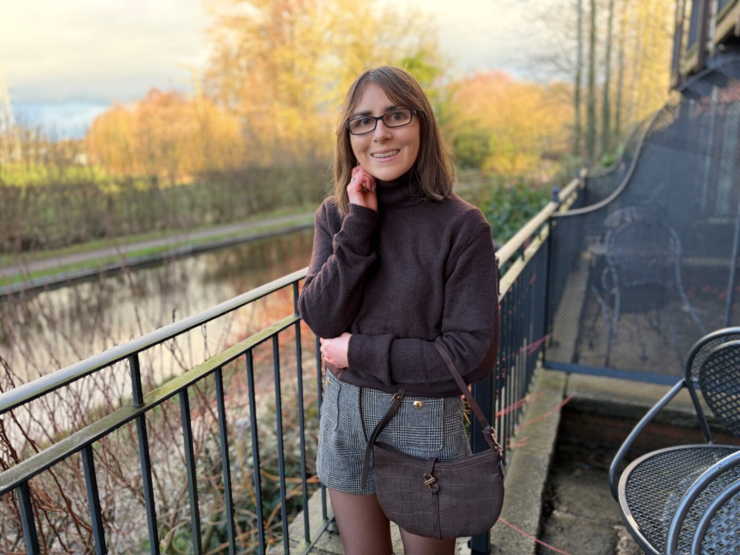 Elin standing on an outdoor terrace beside a canal. She wears a brown high neck jumper with grey and brown check shorts and light brown tights. She’s smiling at the camera with her right hand resting under her chin and her left arm folded across her body. A brown bag hangs in the crook of her left arm. The water beside her reflects the light and there are trees with light orange leaves.