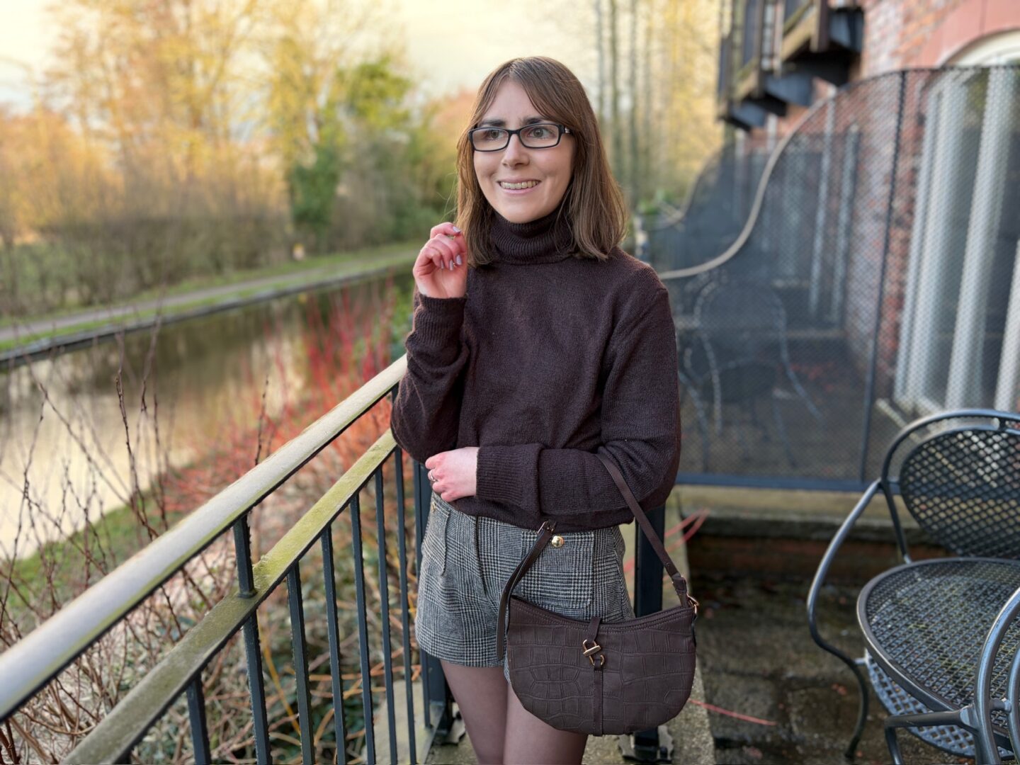 Elin standing on an outdoor terrace by a canal. Her right hand is raised towards her face with her fingers slightly curled inwards, showing her lilac painted nails, and her left arm is across her stomach, acting as a place for her right elbow to rest on. She’s looking away from the camera to her right with a bright smile on her face. She wears a brown high neck jumper with high waisted grey and brown check shorts and a brown shoulder bag which hangs in the crook of her left elbow. Her shoulder length brown hair is worn straight and she has black glasses. The water reflects the trees that line one side.