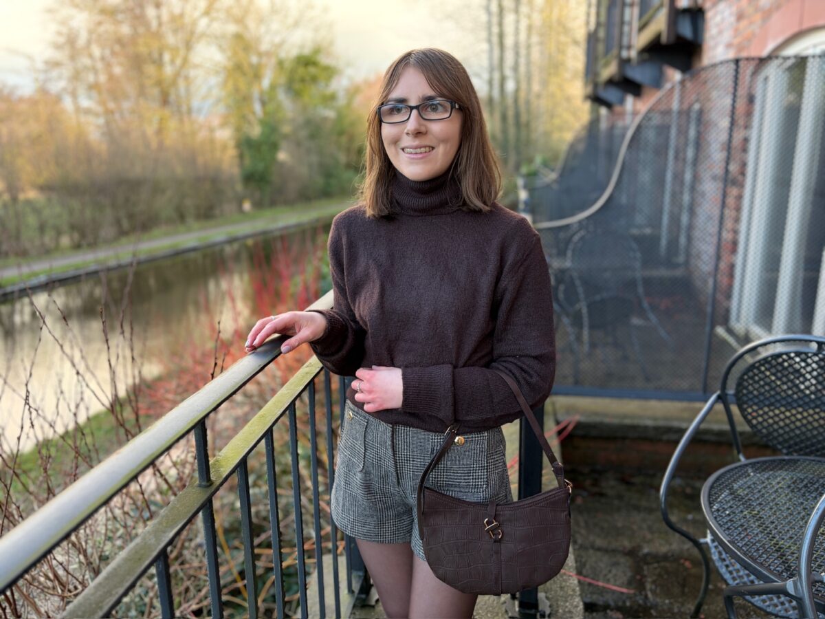 Elin standing on an outdoor terrace by a canal. Her right hand rests on a black railing with two silver rings adorning her fingers. She’s looking away from the camera to her right with a bright smile on her face. She wears a brown high neck jumper with high waisted grey and brown check shorts and a brown shoulder bag which hangs in the crook of her left elbow. Her shoulder length brown hair is worn straight and she has black glasses. The water reflects the trees that line one side.
