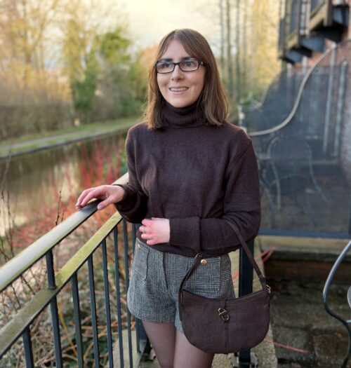 Elin standing on an outdoor terrace by a canal. Her right hand rests on a black railing with two silver rings adorning her fingers. She’s looking away from the camera to her right with a bright smile on her face. She wears a brown high neck jumper with high waisted grey and brown check shorts and a brown shoulder bag which hangs in the crook of her left elbow. Her shoulder length brown hair is worn straight and she has black glasses. The water reflects the trees that line one side.