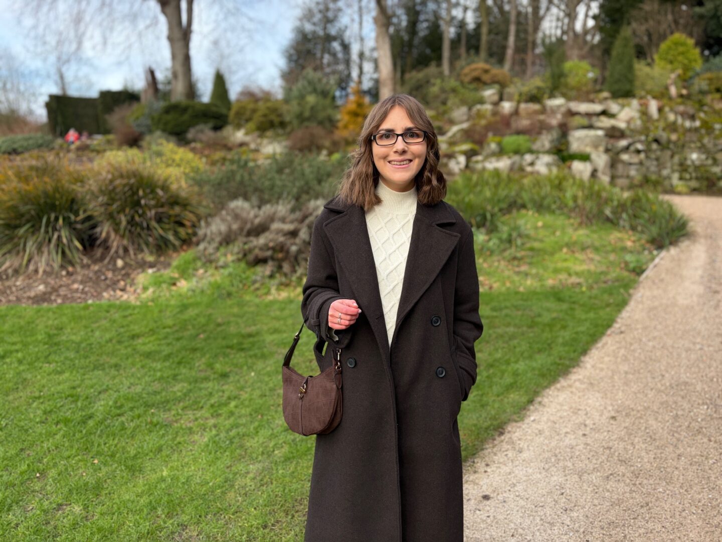 Elin standing on a gravel path that curves through a garden. There is a stone wall behind her with green plants on terraces. She wears a double breasted brown coat over a cream jumper and skirt coord and a brown bag with a croc design hangs on her right arm. Her brown shoulder length hair is styled into loose waves and she’s smiling at the camera.