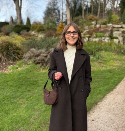 Elin standing on a gravel path that curves through a garden. There is a stone wall behind her with green plants on terraces. She wears a double breasted brown coat over a cream jumper and skirt coord and a brown bag with a croc design hangs on her right arm. Her brown shoulder length hair is styled into loose waves and she’s smiling at the camera.