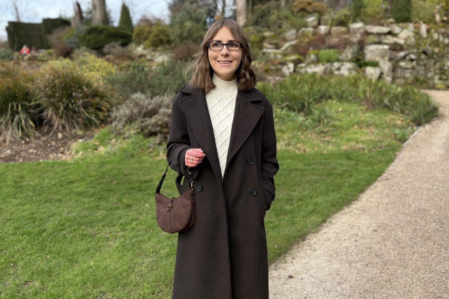 Elin standing in a garden with plants behind her on a stone wall. She’s smiling and looking away from the camera to her right. She wears a long double breasted brown coat over a cream high neck jumper and skirt coord, a brown bag and black glasses.
