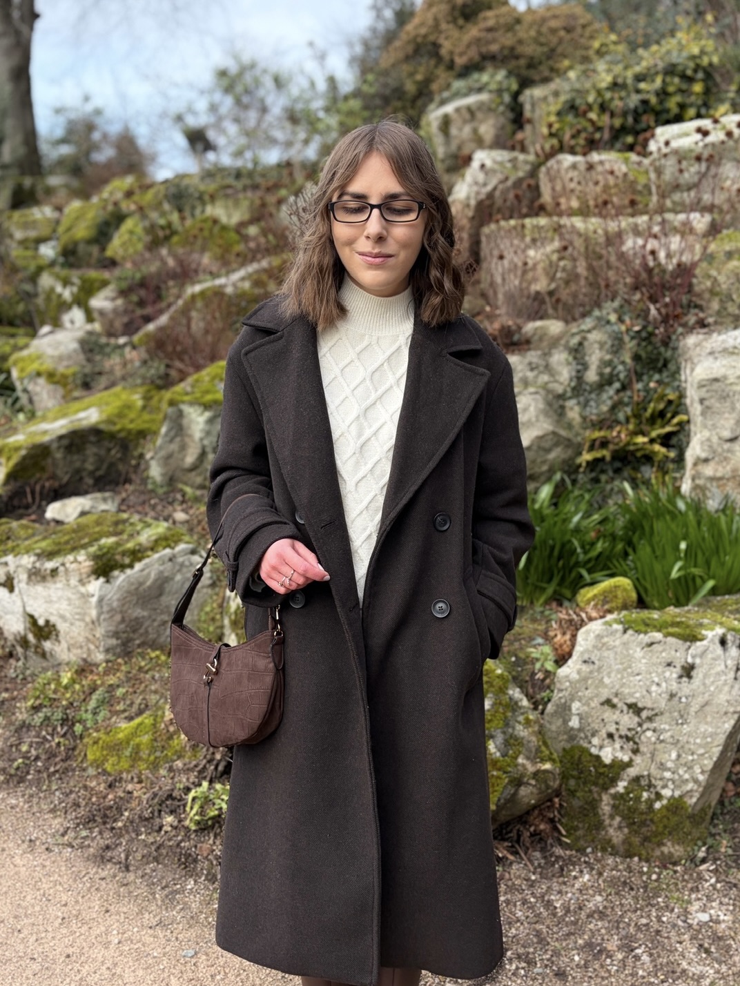 Elin standing in a rocky garden area with large stones, patches of green moss and plants. She wears a long brown coat over a cream jumper and skirt coord and a brown shoulder bag hangs from the crook of her right arm. She’s looking down.