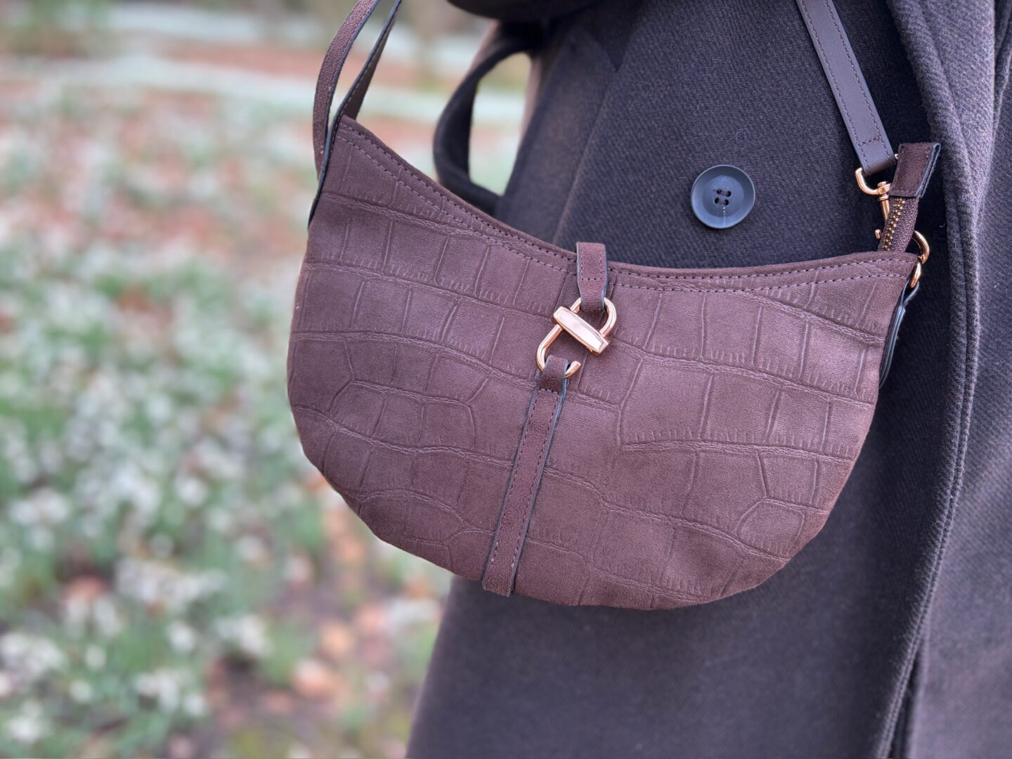A close up shot of a crescent shaped brown handbag against a dark brown coat. The bag has an embossed croc style pattern and has a thin vertical strap on the front that loops through a metal ring and bar detail clasp. A blurry glimpse of the background shows white snowdrop flowers on the ground.