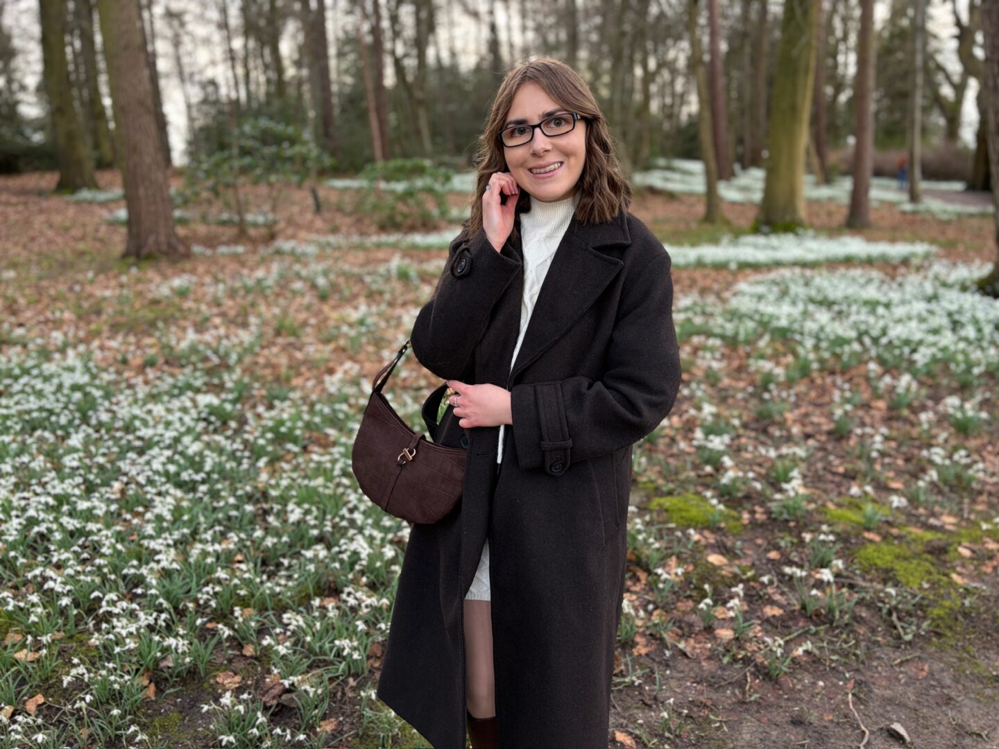 Elin standing in woodland with a carpet of white snowdrop flowers on the ground behind her. She’s smiling broadly at the camera with her right hand raised to just under her chin and the other loosely holding the strap of her brown bag. Her shoulder length brown hair is styled into loose curls and she wears a brown coat over a cream jumper and skirt coord, brown knee high boots and black framed glasses.