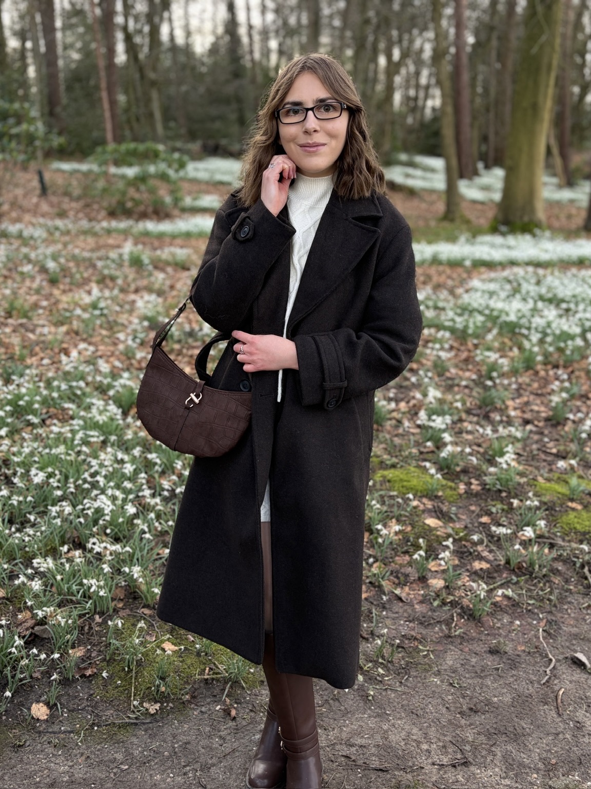 Elin standing on a woodland path which snakes through grass carpeted with white snowdrop flowers. Her right arm is raised to just under her chin and she’s smiling a close lipped smile at the camera.