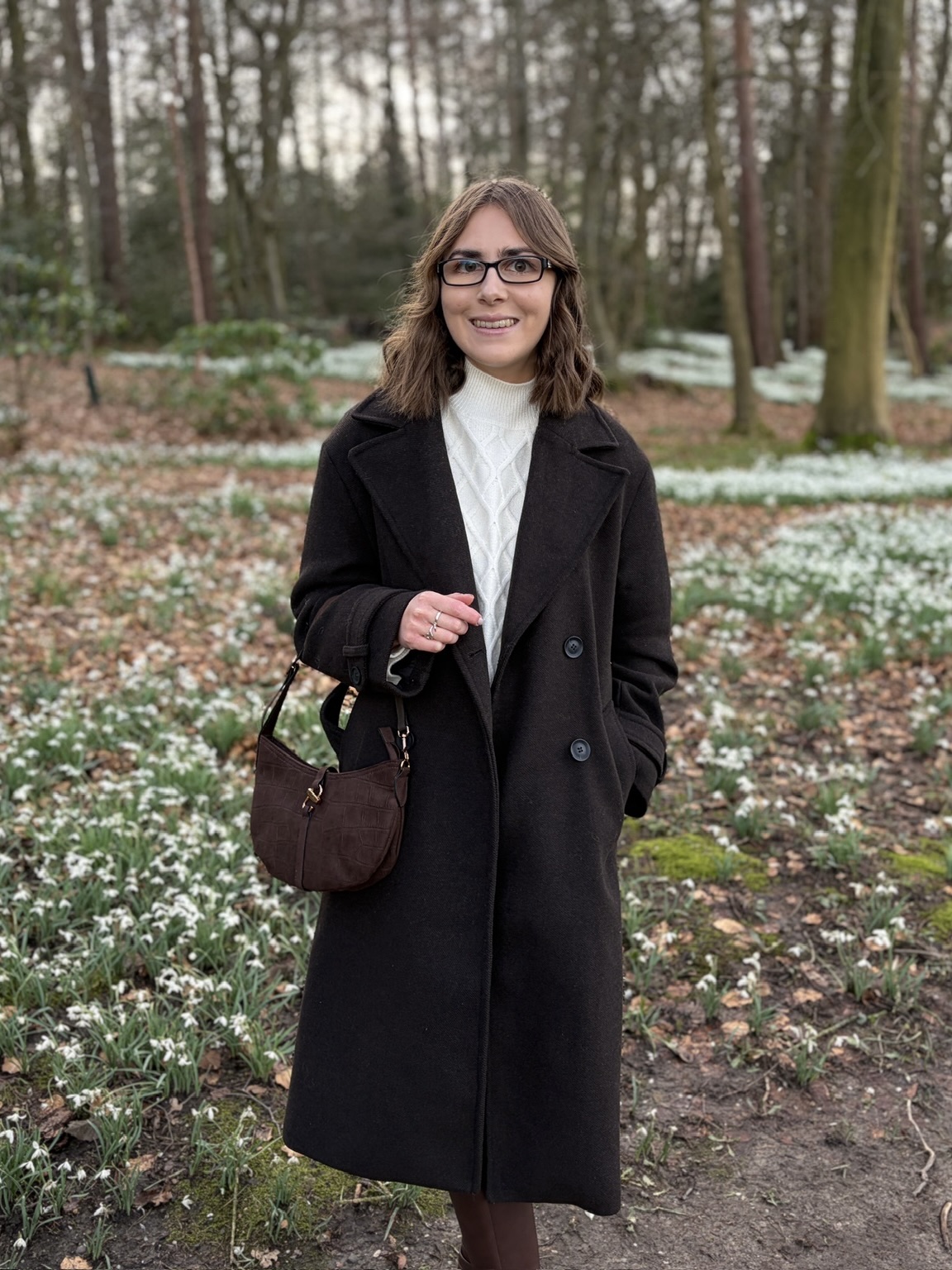 Elin standing in woodland in front of countless snowdrop flowers. She wears a brown double breasted coat over a cream jumper and skirt coord and a brown bag hangs on her right arm. She’s smiling at the camera.