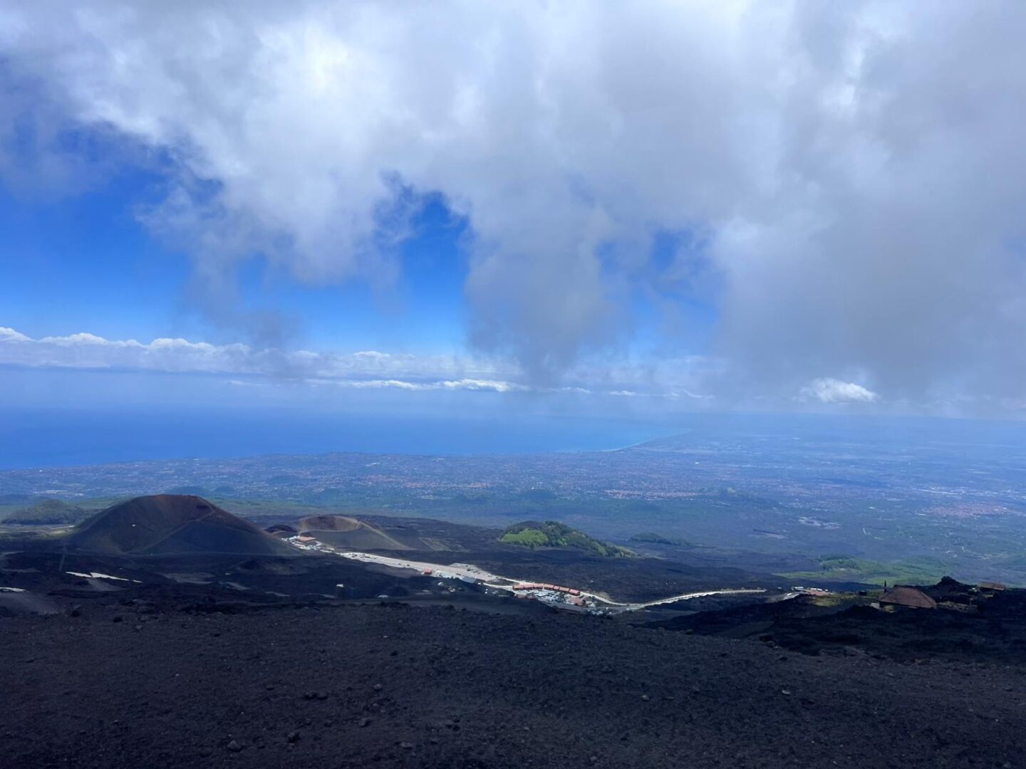 The dark volcanic landscape of Mount Etna stretching towards the coastline under a cloudy sky. The ground is covered in volcanic rock with sparse patches of lighter areas. The view looks out over Catania which has clusters of buildings with red roofs. There are green and brown hills in the distance.