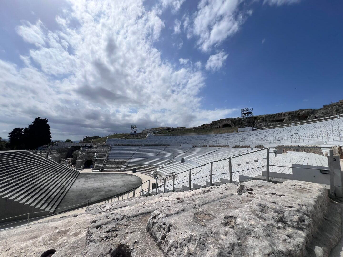 An ancient stone amphitheatre with wide curving seats under a partly cloudy sky. The seating is set out in a semi circle stepping upwards in many tiers. At the bottom is a dark stage area.