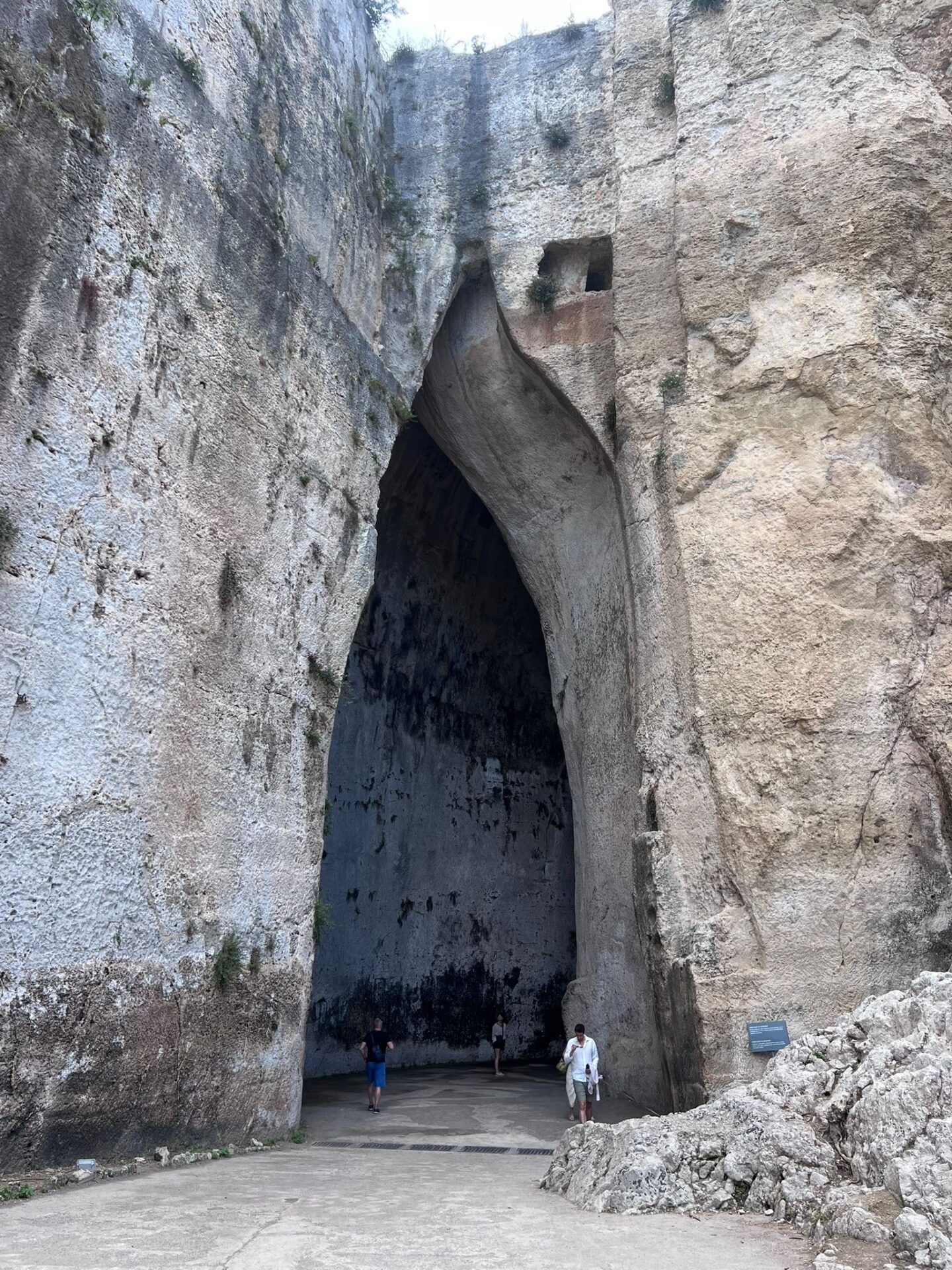 A massive stone cave entrance in a steep rock face. The opening is tall and narrow, tapering upward with a curved, ear shaped, shape.