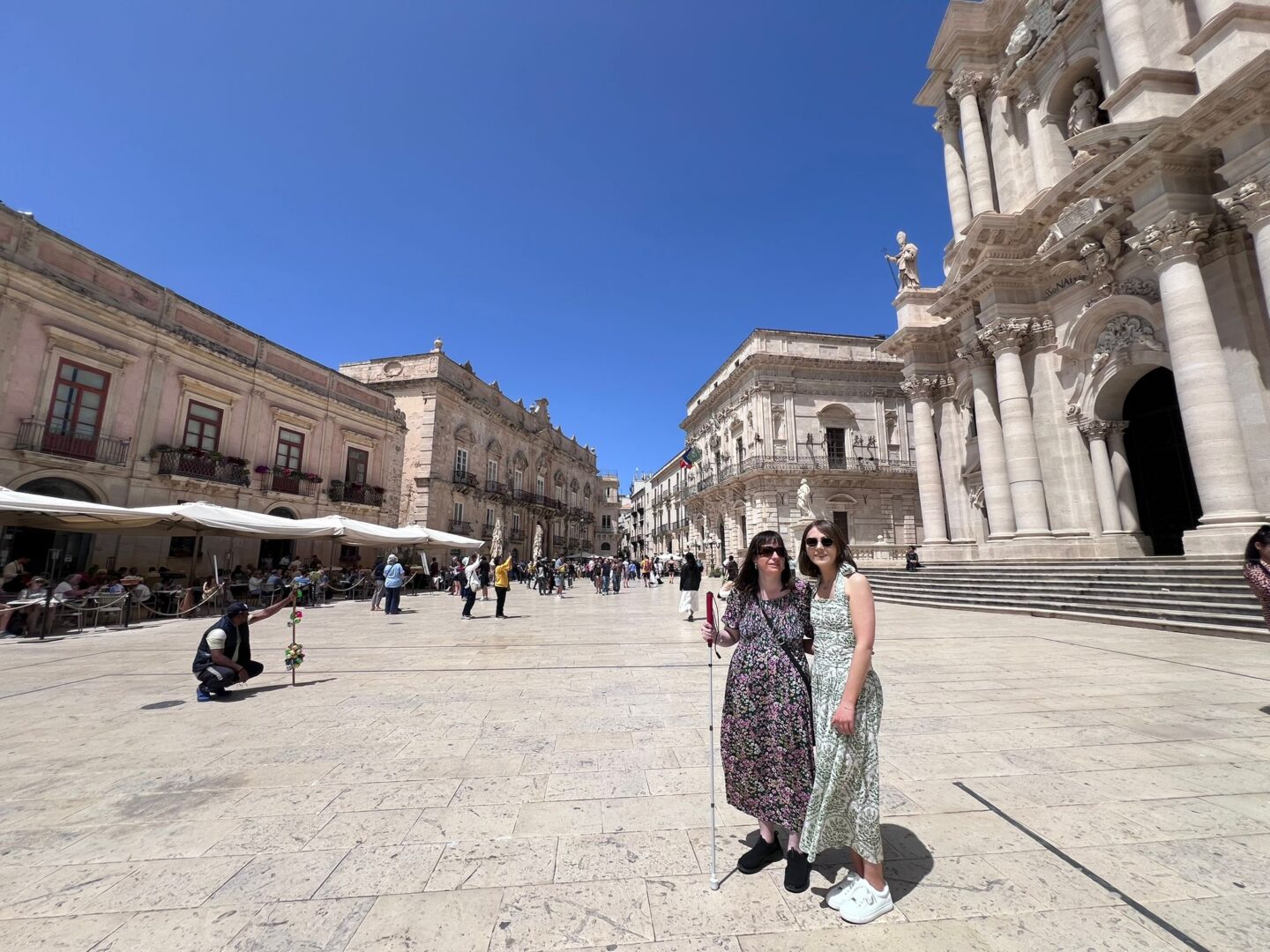Elin and Holly standing together in Ortigia's large sunlit square. Grand historic buildings frame the area and they're standing in front of an ornate cathedral with tall columns, statues and a wide staircase. Holly wears a black floral dress and black trainers with a cane in her hand and Elin wears a white maxi dress with a halterneck and green floral details with white trainers. They're both wearing sunglasses.
