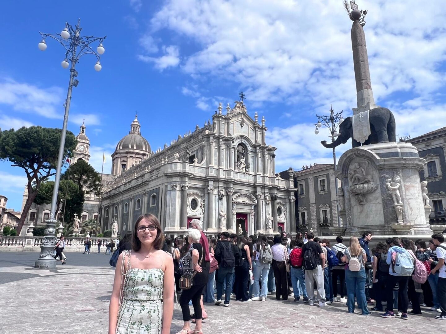 Elin standing in Catania's main square. She wears a coord set which looks like a dress, it features a top with thin spaghetti straps, a fitted bodice, pleats in the top and skirt and a green vine design. Behind her, a large group of people are gathered by a tall fountain with an elephant on top. To the left, a grand baroque style cathedral features statues, ornate carvings and a large central dome. Several walls are draped with dark red fabric.