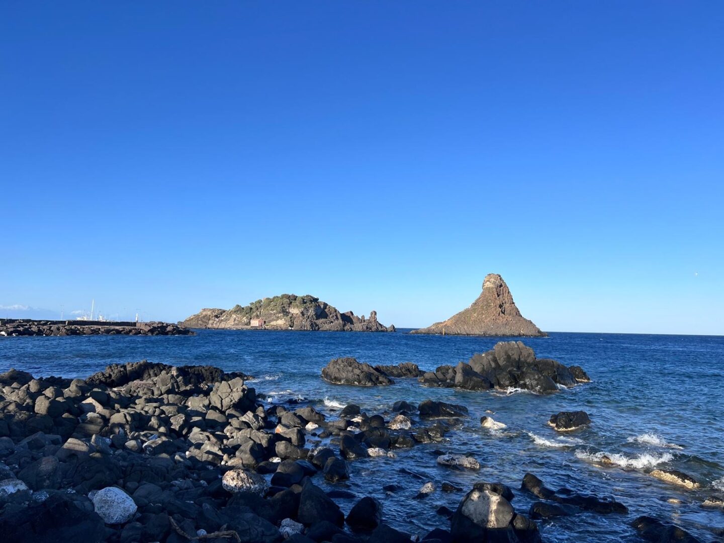 A rocky shoreline beside a deep blue sea. In the distance, several rock formations rise up from the water looking irregular in shape. To the left, a rocky island has patches of green vegetation and a small light coloured building near the shore. The sky is bright and cloudless.