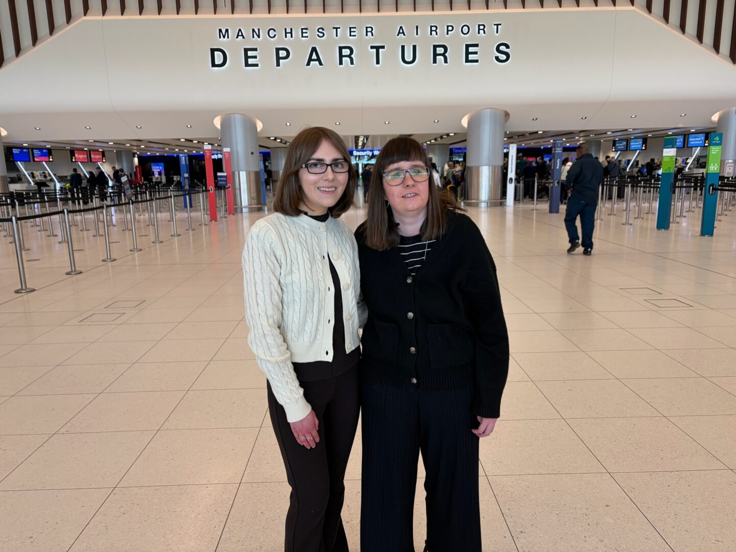 Elin and Holly standing with their arms around each other in front of the Departures sign at Manchester Airport. They're both smiling broadly at the camera, excited about their trip.