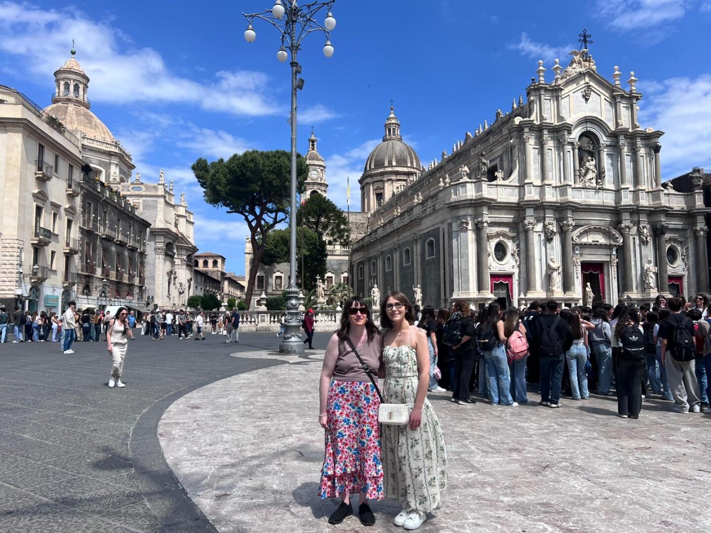 Elin and Holly standing in Catania's main square in front of an ornate cathedral. It features columns, statues and large domes. They have their arms around each other and are smiling at the camera. Holly wears a mauve sleeveless top with a long colourful skirt, black trainers and a black crossbody bag. Elin wears a coord set which features a pleated top and maxi skirt with a green vine design. She's paired it with a white handbag and white trainers.
