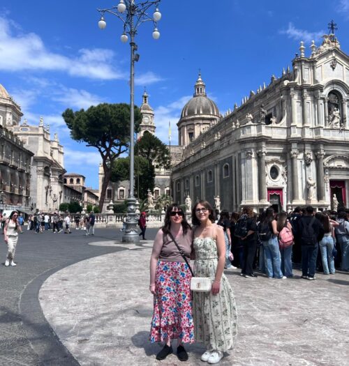 Elin and Holly standing in Catania's main square in front of an ornate cathedral. It features columns, statues and large domes. They have their arms around each other and are smiling at the camera. Holly wears a mauve sleeveless top with a long colourful skirt, black trainers and a black crossbody bag. Elin wears a coord set which features a pleated top and maxi skirt with a green vine design. She's paired it with a white handbag and white trainers.