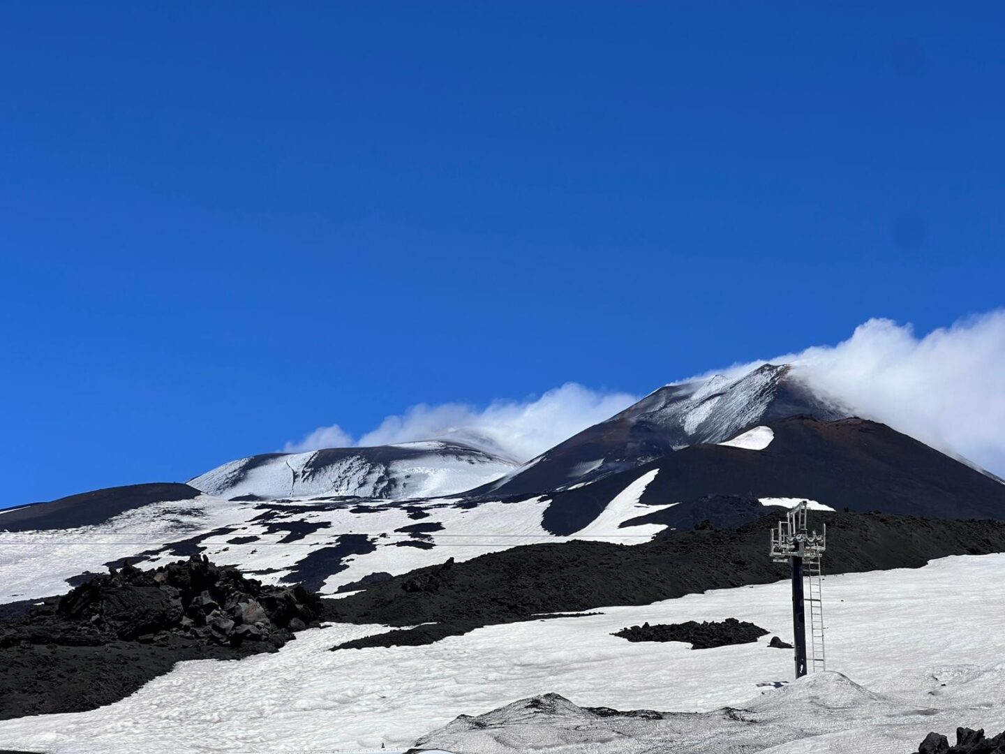 Snow covered volcanic slopes under a bright blue sky. The snow is broken up by dark jagged lava rocks.