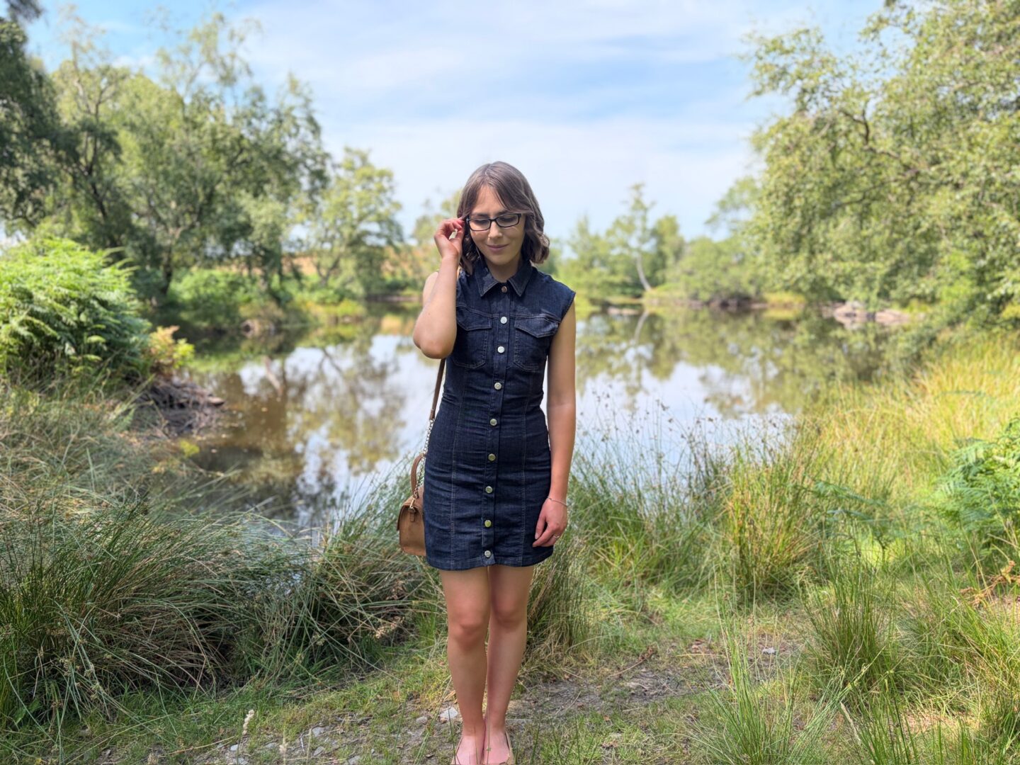 Elin standing in front of a small lake which is bordered by trees. She wears a dark denim mini dress with tan accessories and she's looking down. Her right arm is raised to hold a strand of hair away from her face.