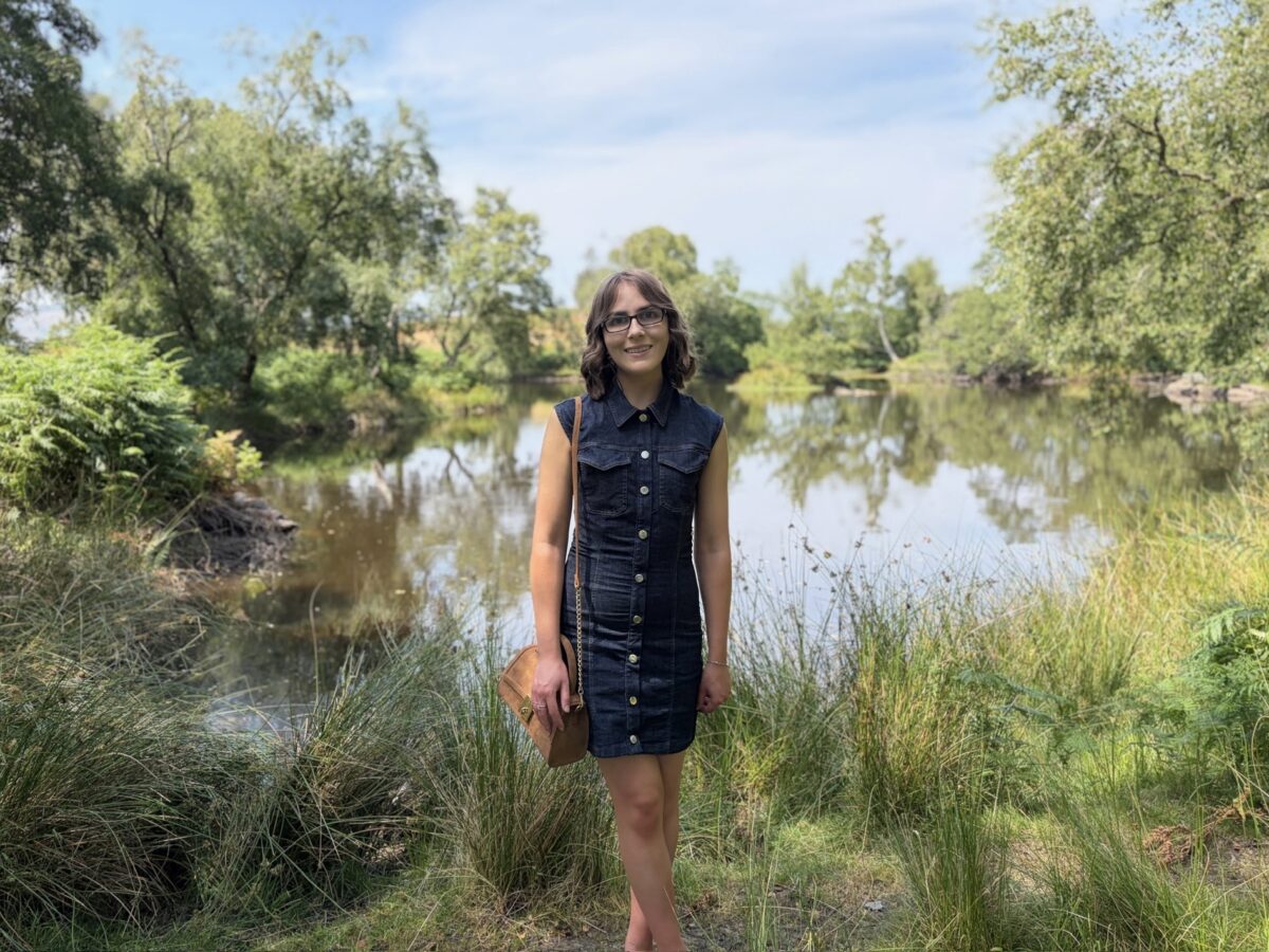 Elin standing in front of a small lake which is bordered by trees. She wears a fitted sleeveless dark denim dress with gold buttons down the centre. It's paired with a tan bag and tan ballet shoes. Her shoulder length brown hair is styled into waves and she's smiling at the camera.