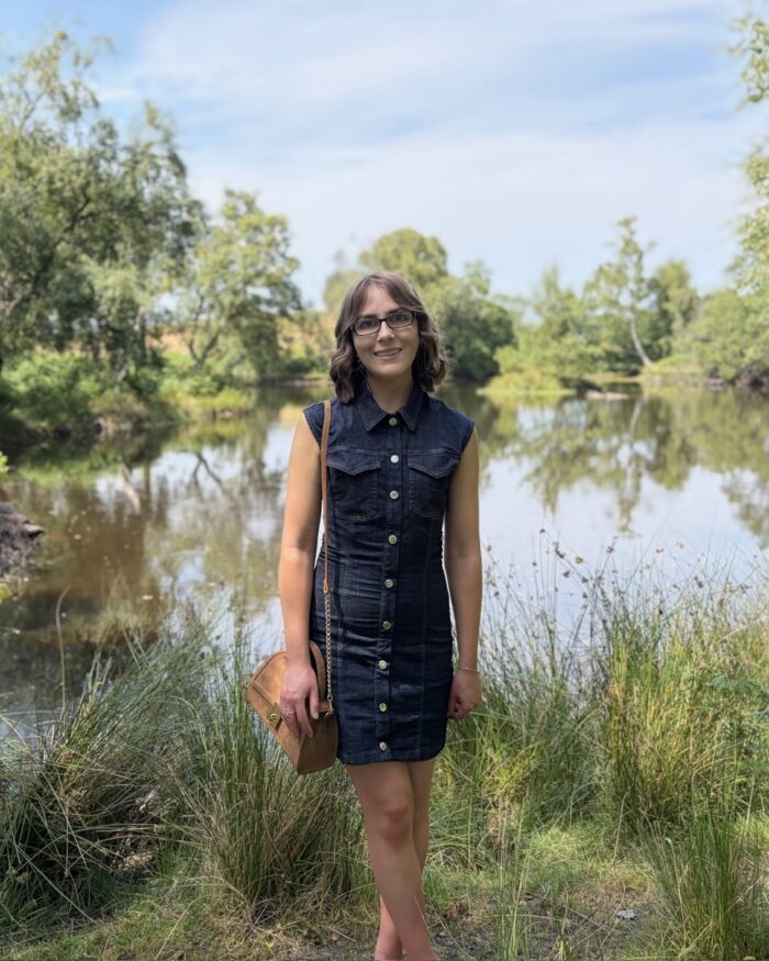 Elin standing in front of a small lake which is bordered by trees. She wears a fitted sleeveless dark denim dress with gold buttons down the centre. It's paired with a tan bag and tan ballet shoes. Her shoulder length brown hair is styled into waves and she's smiling at the camera.