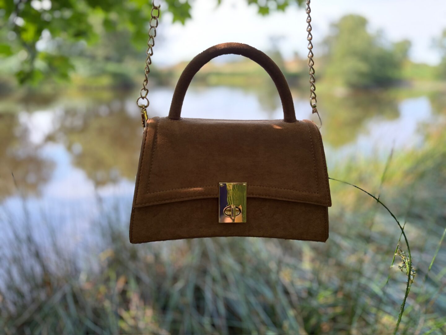 A tan coloured handbag hanging from a tree branch which is out of frame. The bag has a gold buckle on the front and a small grab handle at the top as well as a long cross body strap. The background is slightly out of focus but shows a small lake surrounded by trees and greenery which are reflected on the water.