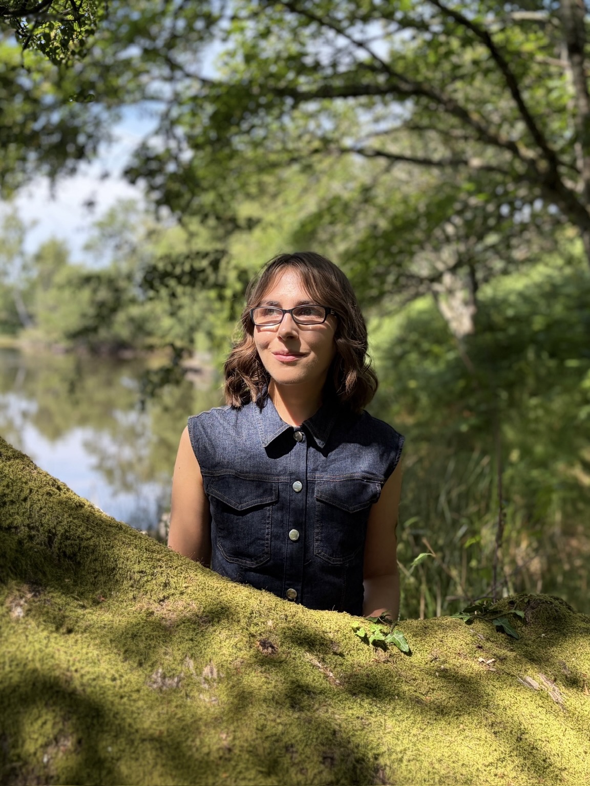 Elin standing in woodland dappled in sunlight by a small lake. The sun casts her in light and shadow and she looks away from the camera with a thoughtful expression.