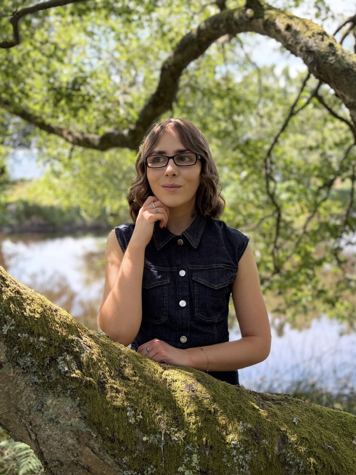 Elin standing in woodland behind a thick tree branch. Her left hand rests on it whilst the other is raised to under her chin as she looks away from the camera. Sunlight filters through the leaves behind her and a small lake is also visible.