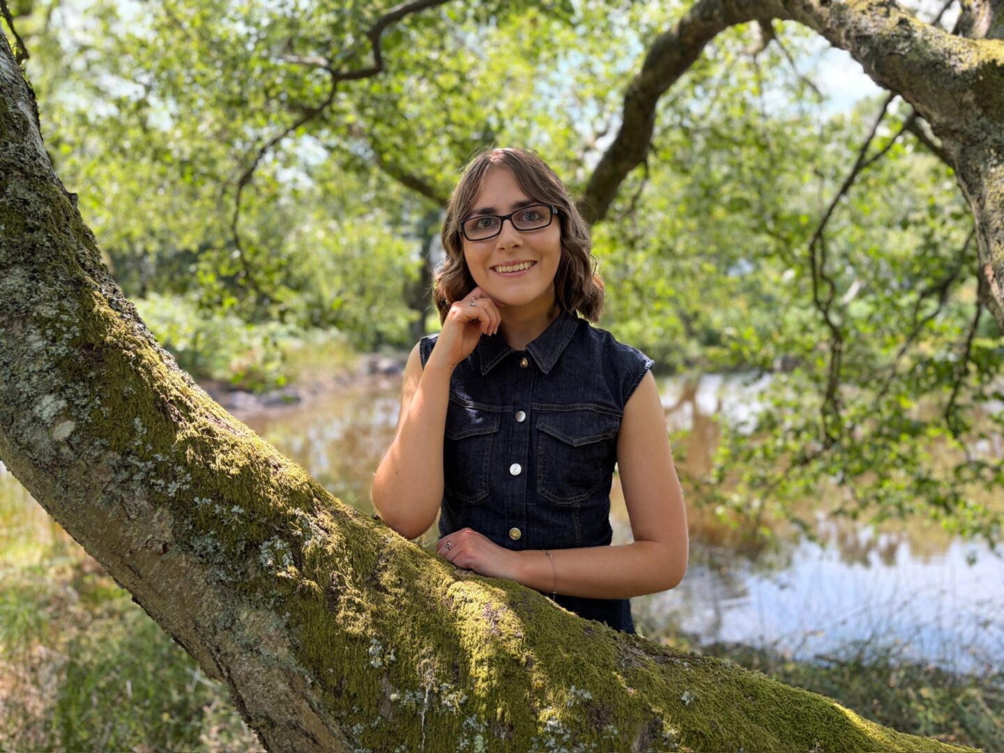 Elin standing in woodland behind a thick tree branch with her left arm resting on it whilst her right arm is raised to just under her chin. She wears a black denim dress with gold buttons down the front and two chest pockets. Her shoulder length brown hair is styled into waves and she's smiling broadly at the camera.