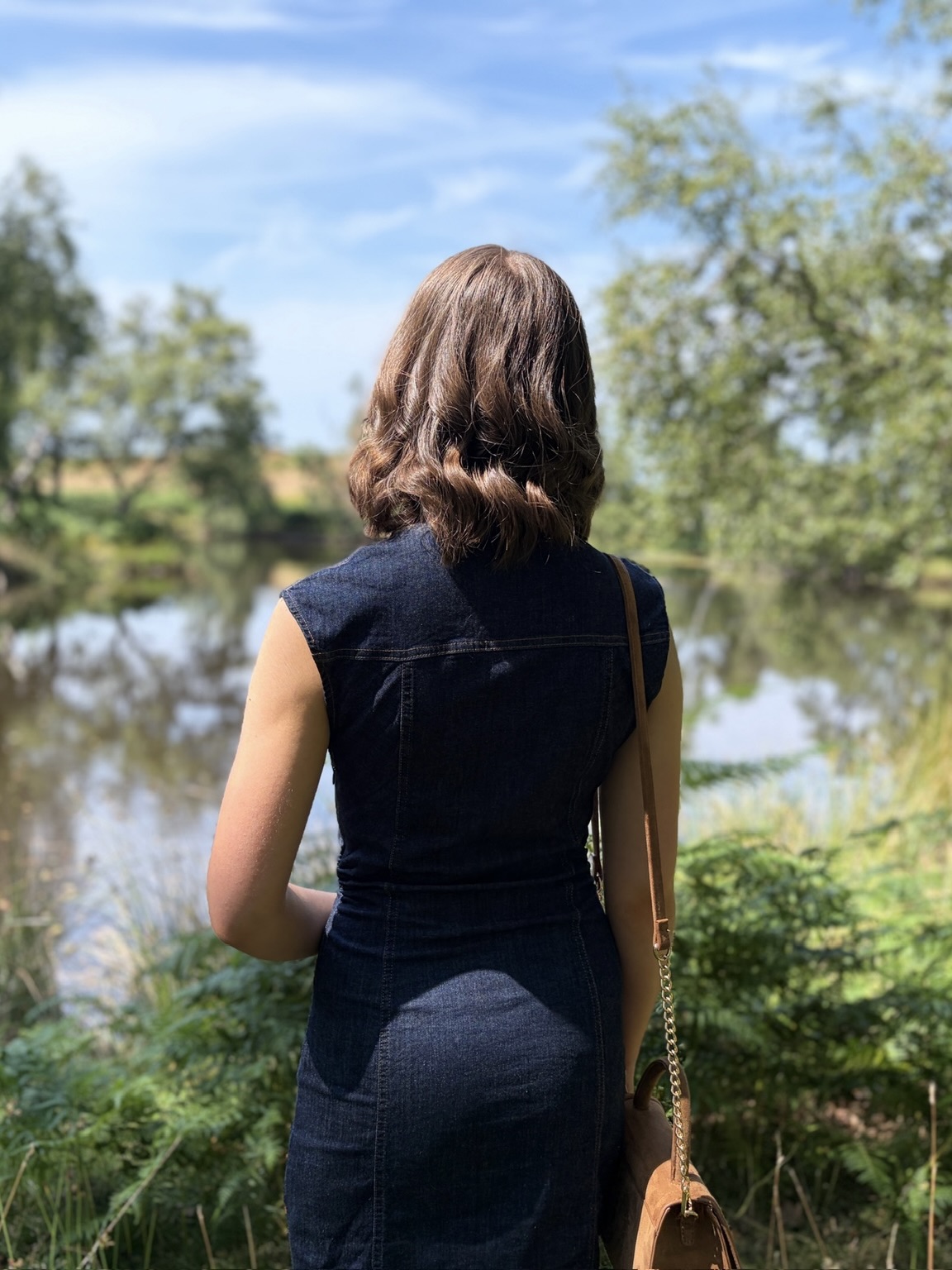 Elin looking out over a calm lake which is bordered by trees. The sun casts her in light and shadow, highlighting her brown wavy hair, and the trees and greenery around the lake are reflected on the water.