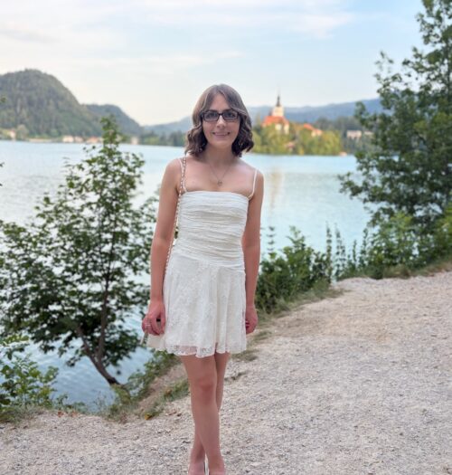 Elin standing on a path surrounding Lake Bled. She's smiling at the camera and wears a white mini dress with delicate lace details all over, a white handbag which has a chain strap, a silver necklace and bracelet and dark framed glasses. Her short hair is styled into soft curls. The lake is bordered by trees and greenery and the water is calm with its surroundings reflected on the surface. The famous island can be seen behind her with the spire of the church reaching towards the sky. Tall mountains can be seen in the distance.