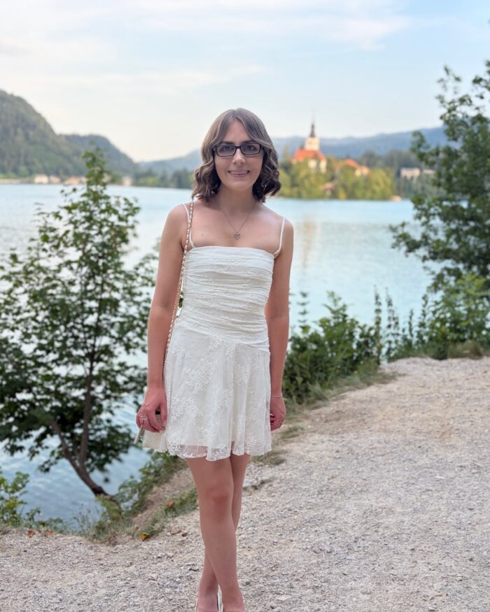 Elin standing on a path surrounding Lake Bled. She's smiling at the camera and wears a white mini dress with delicate lace details all over, a white handbag which has a chain strap, a silver necklace and bracelet and dark framed glasses. Her short hair is styled into soft curls. The lake is bordered by trees and greenery and the water is calm with its surroundings reflected on the surface. The famous island can be seen behind her with the spire of the church reaching towards the sky. Tall mountains can be seen in the distance.