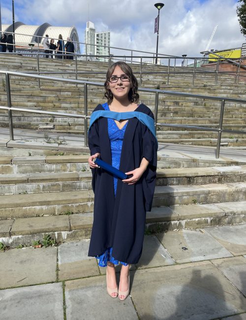 Elin in her graduation gown which is navy with a light blue and yellow hood. It's worn over a blue lace midi dress and she's holding a dark blue cylinder in her hands. Her brown hair reaches just above her shoulders and it has been curled and styled into a half-up, half-down do. The sun is shining on Elin as she smiles at the camera, standing in front of stone steps that lead up to the Bridgewater Hall.