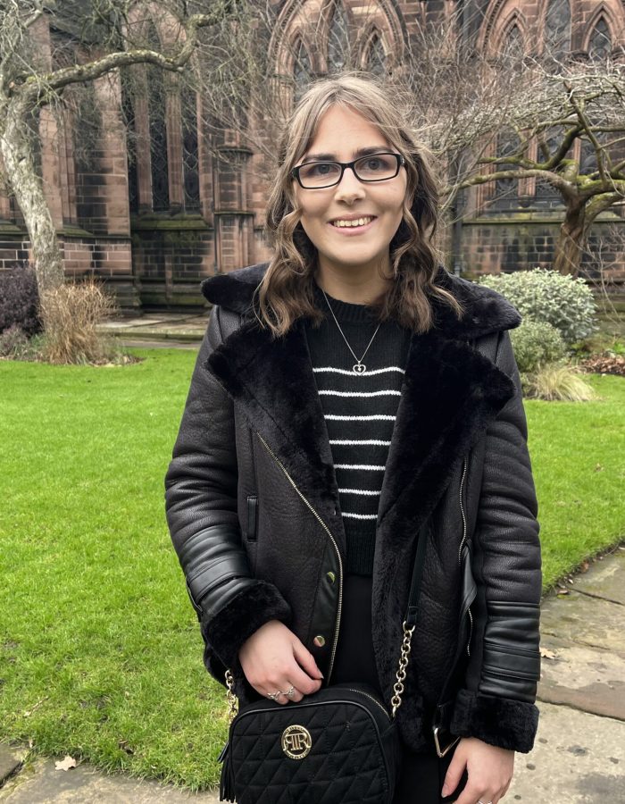 Elin standing on a stone path in front of a cathedral which has large gothic style windows. The path is bordered by a manicured lawn. She wears a black aviator jacket which has faux fur lining and a belt, it's worn open over a black and white striped crop jumper and a black skort. The outfit is accessorised with a black cross body bag which has gold hardware and silver jewellery including three rings and a dainty heart shaped necklace. Elin is smiling at the camera.