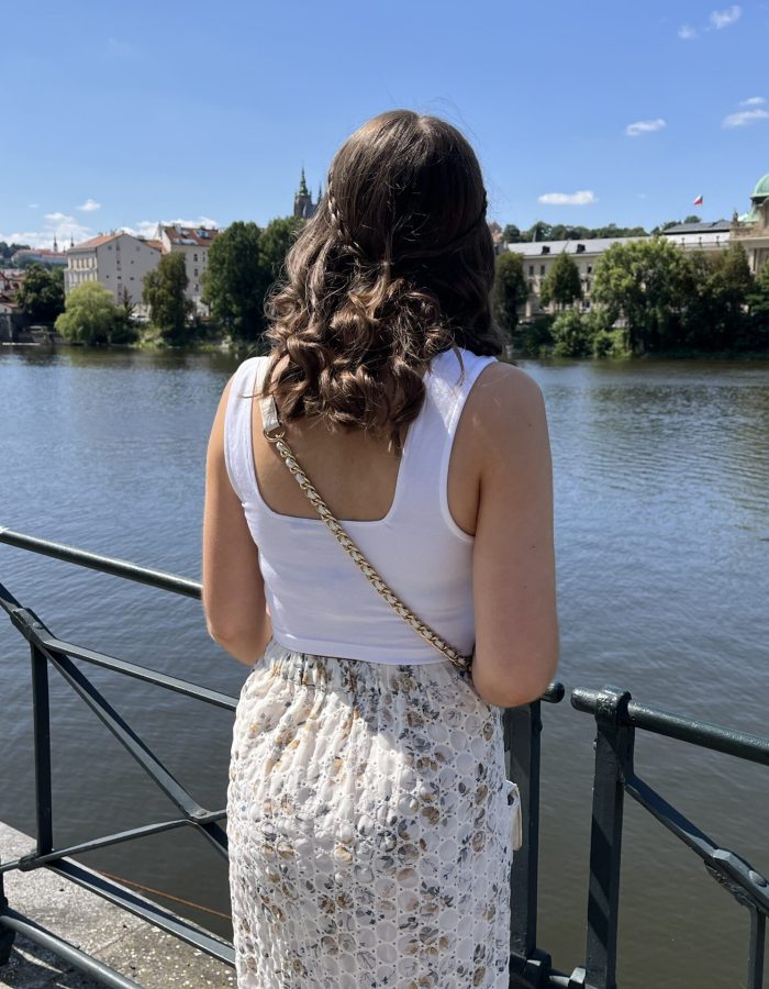 Elin looks out over the Vltava river in Prague on a sunny day. Her hair is styled into a curly half-up, half-down do with two small braids wrapping around her head to meet in the middle. She wears a white crop top which has wide straps, a beige broderie midi skirt and a white crossbody bag.