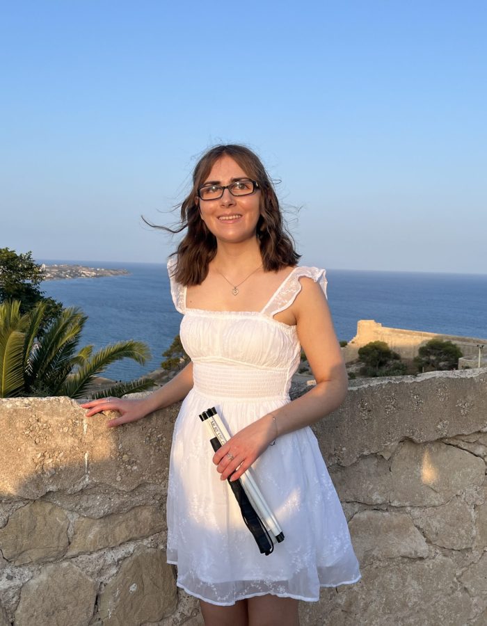 Elin standing by a stone wall with her right hand resting on it. She wears a white mini dress with embroidered details, a square neckline and sheer flutter sleeves. She holds her white cane in her left hand. A mix of scenery is in the background from the tall buildings of a city to greenery and the sea.
