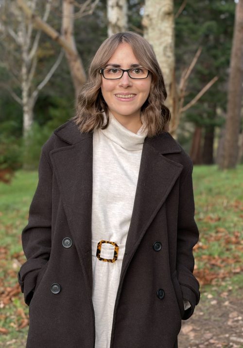 Elin standing in the woods with fallen leaves on the ground all around her. She's wearing a chocolate brown double breasted coat over a cream belted jumper dress which has a roll neck. Her hands are in her coat pockets and she's smiling broadly at the camera. Her brown hair is styled into loose waves which hang just above her shoulders.