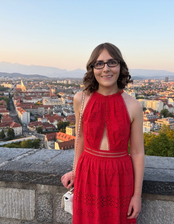 Elin standing in front of a stone wall overlooking a cityscape. She wears a bright red maxi dress which has a halter neck and broderie anglaise details all over. It has a triangle shaped cutout at the waist. Her short brown hair is styled into soft curls and she wears glasses. A white bag hangs from her right shoulder with a chain strap and her right hand rests on the wall. She's smiling broadly at the camera.