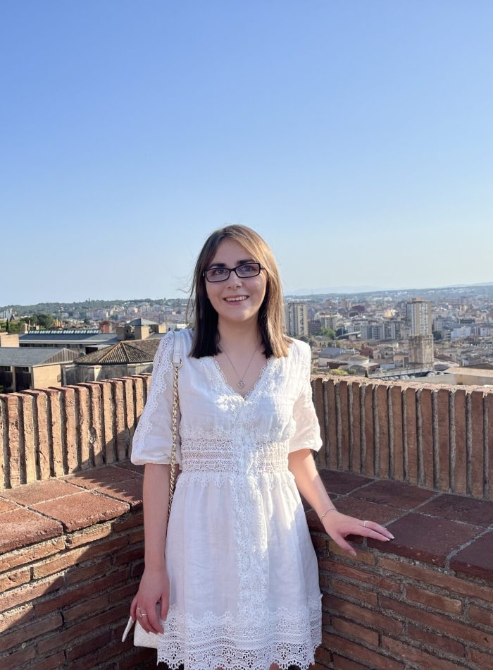 Elin standing with her hand on a stone wall which overlooks the city of Girona in Spain. She wears a white mini skater dress which has lace detailing around the waist, down the centre, along the hem and on the sleeves which fall just below her elbows. It’s paired with a white handbag which has gold hardware. Her shoulder length brown hair is worn straight and she’s smiling broadly at the camera.